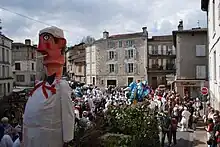 En premier plan, le mannequin Bufador et le bûcher sont remorqués par un tracteur sur une place de Nontron. Les Soufflaculs et les spectateurs sont situés tout autour.