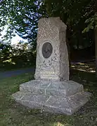 Photographie en couleurs d'une sculpture en forme de menhir, arborant un médaillon en son centre, placée sur une pelouse sous un grand arbre.