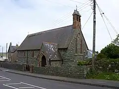 Photo d'un bâtiment allongé aux murs gris et au toit en ardoises, avec un clocher en briques, au bord d'une route dont il est séparé par un mur bas