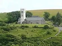 Photo d'un bâtiment allongé aux murs gris et au toit en ardoise doté d'une grande tour carrée blanche sur la gauche, au cœur d'un paysage verdoyant
