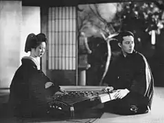 Une jeune femme (Kinuyo Tanaka) en kimino sombre est agenouillée sur le tatami d'une pièce traditionnelle japonaise dont la porte coulissante s'ouvre sur un jardin. Elle joue du koto. Un jeune homme (Kōkichi Takada) agenouillé face à elle tient dans ses mains un paquet et regarde vers la droite par dessus son épaule (image en noir et blanc).