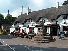Photo d'une grande croix en pierre sur un piédestal de quelques marches devant une grande maison aux murs blancs