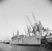 Photographie en noir et blanc d'un navire amarré à un quai, à côté de grues métalliques.