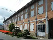 Photo couleur de la façade d'un entrepôt à deux étages, construction en briques rouges aux volets blancs fermés. Devant le bâtiment : un arrangement floral fait d'arbustes au feuillage vert et de massifs de fleurs rouges. Un ciel nuageux en arrière-plan.