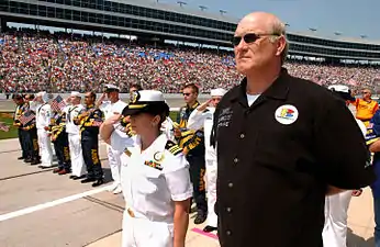 Un homme imposant se tient droit, debout, devant des militaires au garde à vous et une foule massée en tribune.