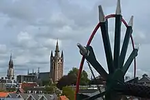 Photographie en couleurs d'un paysage urbain vu en hauteur, un clocher d'église pointant sur la gauche, une roue peinte en noir rouge et blanc et munie de trois axes visible sur la droite.