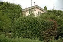 Photo en couleur d'un bout de façade claire sous un toit pointu émergeant de massifs d'arbres et de verdure.