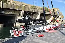 Vu de trois quarts arrière, voilier rouge, blanc et gris amarré au ponton, devant la base de sous-marins de Lorient.