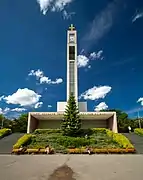 Une église moderne avec en son centre un impressionnant clocher parallélépipédique surmonté d'une croix d'or ; le corps de l'édifice monte en escalier le long de la pente du terrain ; de la pelouse et des fleurs au premier plan.