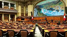 Photographie d'une grande salle munie de pupitres et de chaises, au fond une tribune et sur la droite en haut une grande peinture représentant un paysage