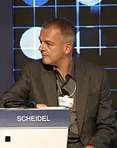 Photographie en buste d'un homme plutôt âgé. Il regarde un individu hors champ et est vêtu d'un costume marron foncé.