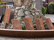 Photographie en couleur montrant un amas de pierres entouré de plusieurs plaques de métal rouillé, gravées avec des noms