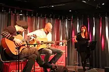 Photo en couleur d'un groupe jouant de la musique dans une pièce, deux guitaristes sont assis à gauche, une chanteuse aux cheveux bruns est debout derrière un pupitre à droite