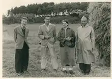 Groupe de quatre personnes dans un champs, avec une botte de foin sur la droite et une forêt en arrière-plan. Jacoba Hol est au centre de la photo.