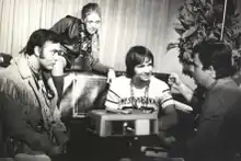 Photographie en noir et blanc de trois hommes assis. L'homme du milieu est assis sur un canapé, une femme est assise sur le dessus de ce meuble et est penchée vers lui.